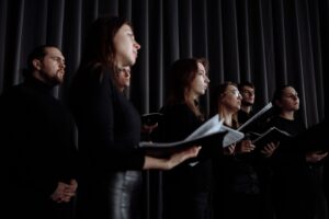Zes zangers met bladmuziek tijdens een kooruitvoering op een podium